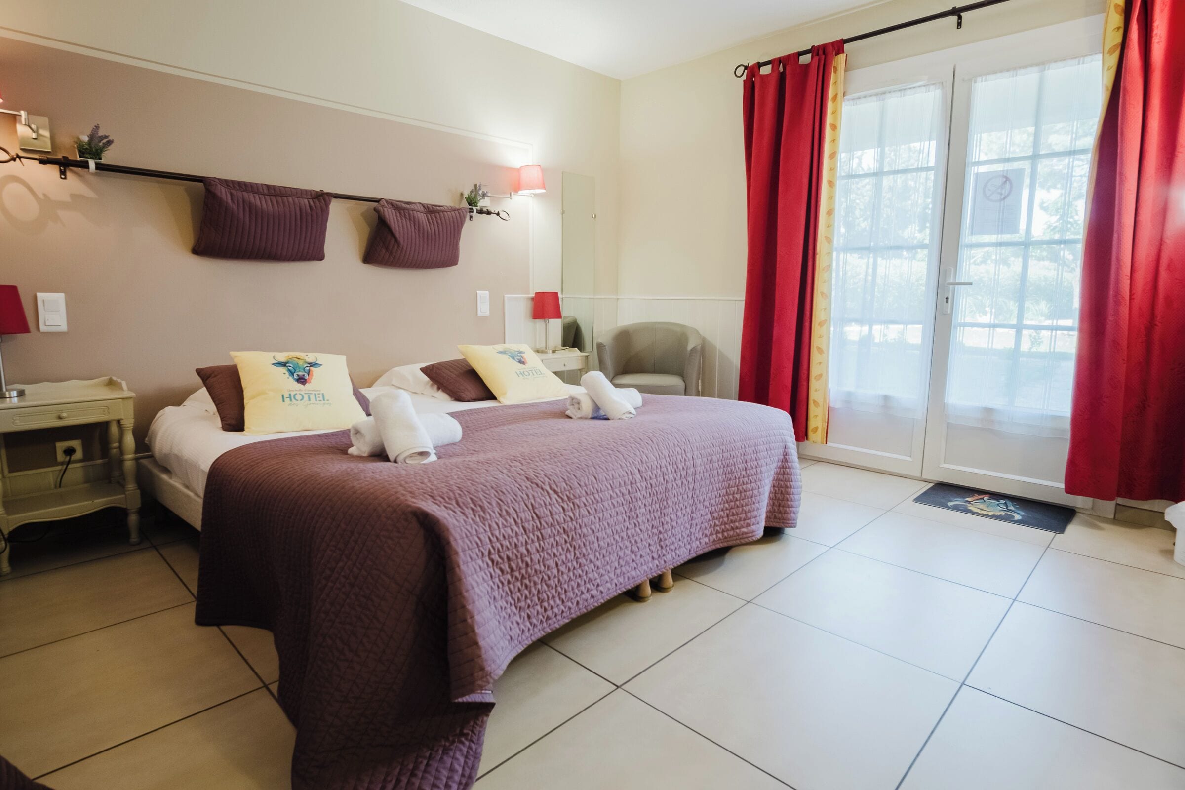 Quarto de hotel com uma cama grande, duas almofadas, mesa de cabeceira, uma cadeira e janela com cortinas vermelhas.