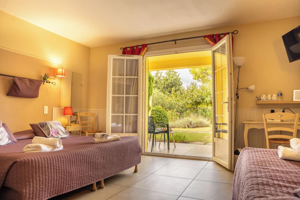 A hotel room with two beds, a desk, a chair, and a sliding glass door leading to a garden.