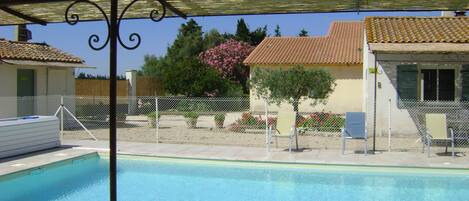 Une piscine entourée d’une clôture en métal, une pergola au-dessus et, au fond, un bâtiment avec un toit en tuiles.