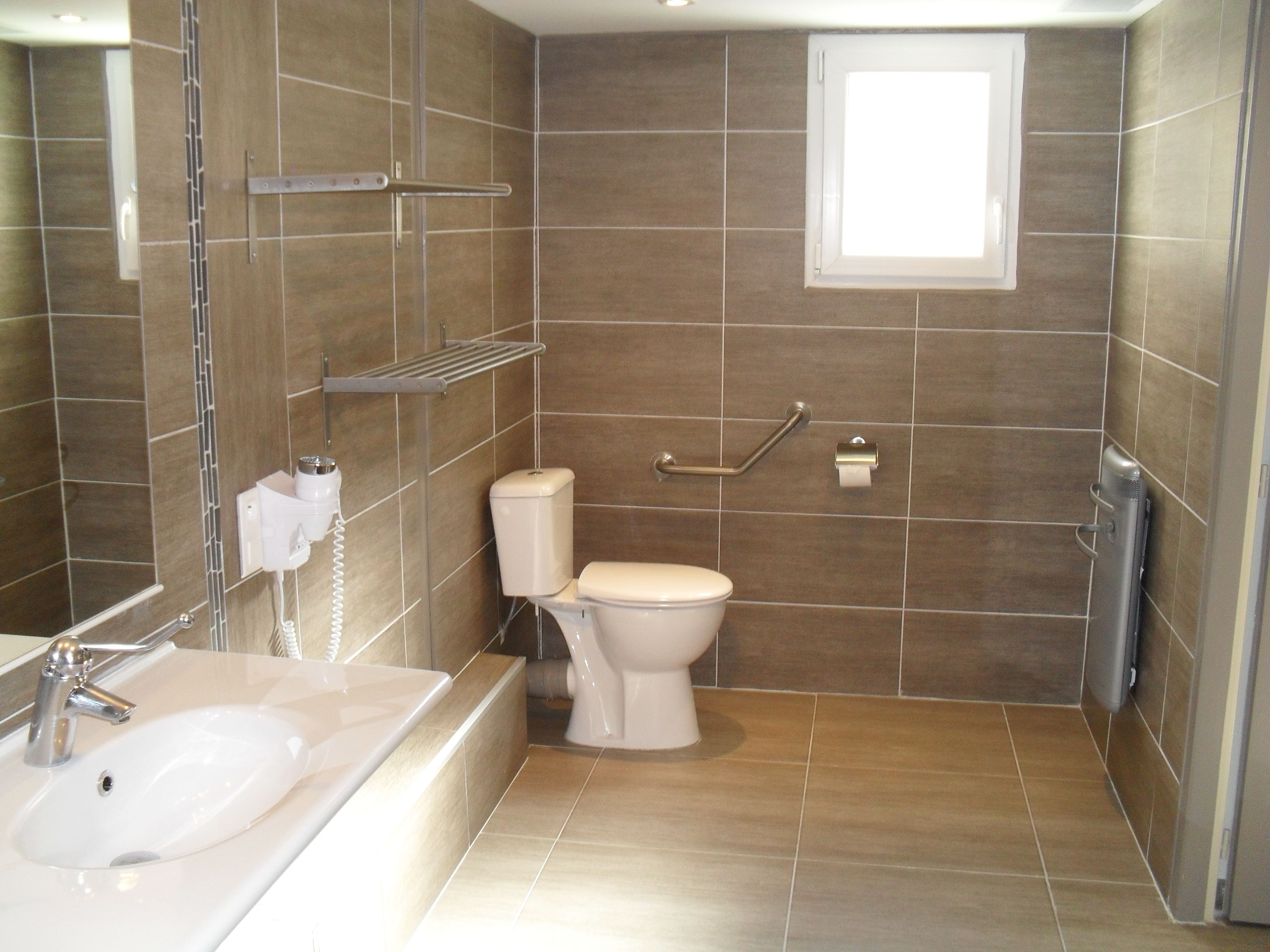 A bathroom with a bathtub, toilet, and a sink. There are grab bars installed near the toilet and a towel dispenser.