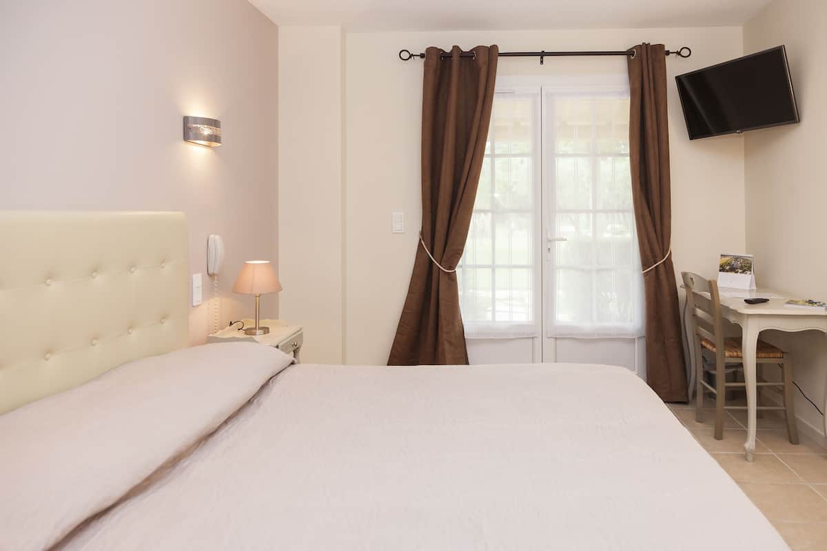 A bedroom with a bed, bedside lamp, window with curtains, a desk with a chair, and a flat-screen TV mounted on the wall.
