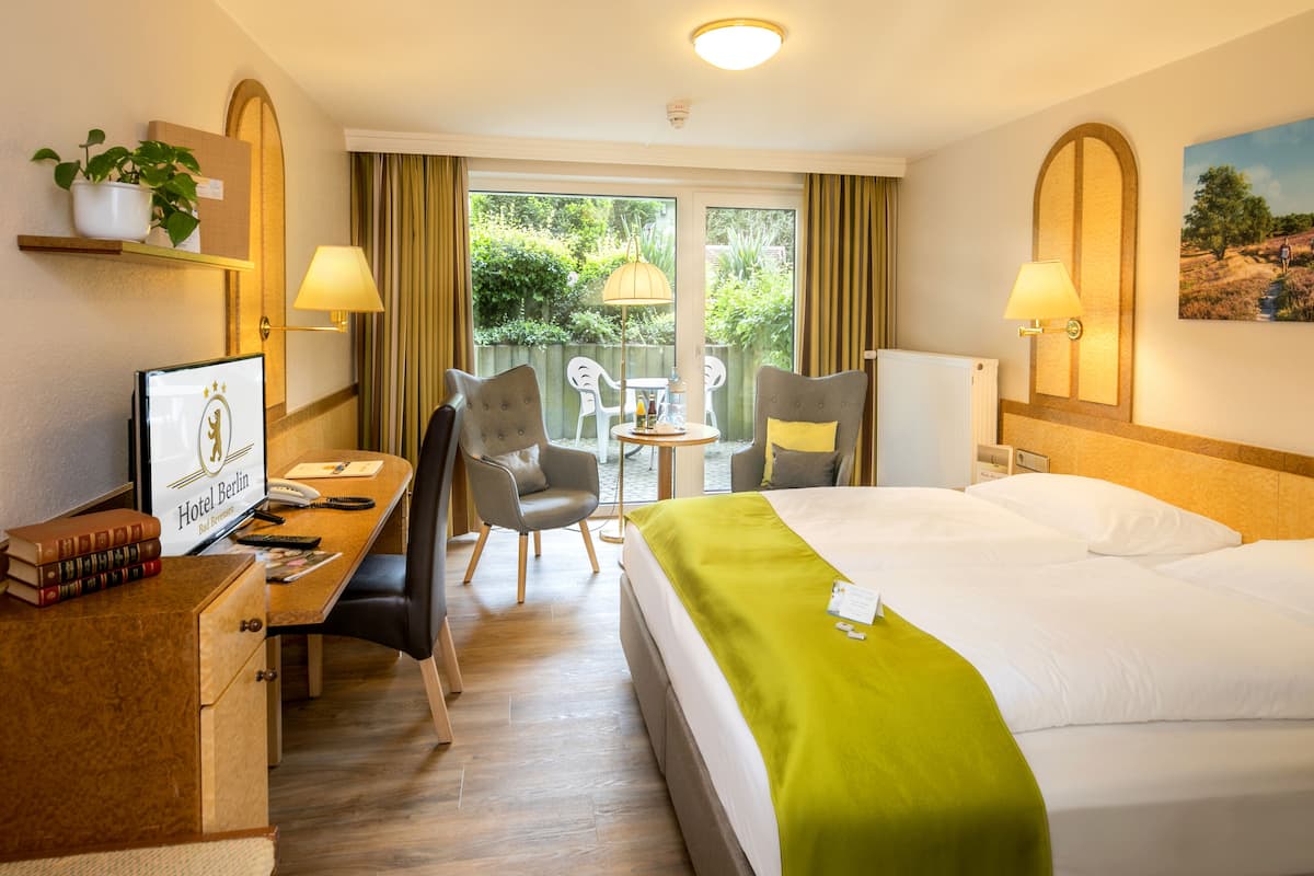 A hotel room with a bed, desk, chairs, a small table, a potted plant, and a painting on the wall.