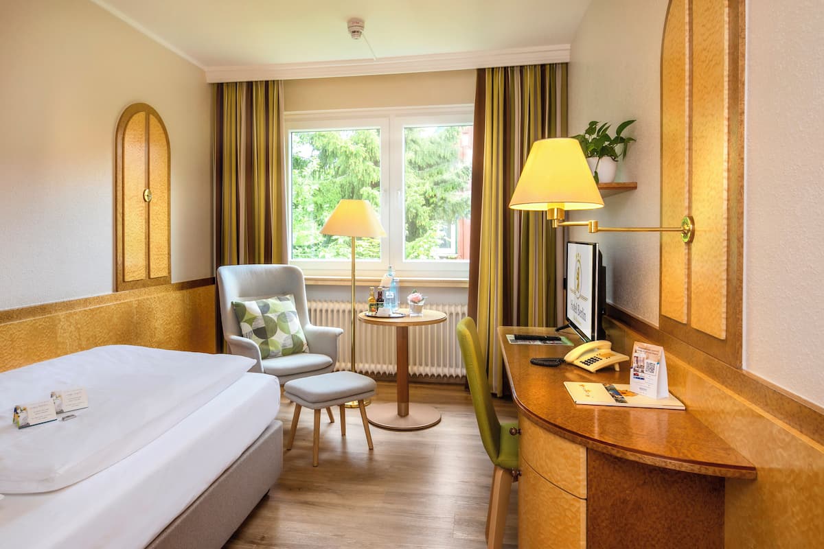 A hotel room with a bed, a desk with a telephone and a small table, a chair, a window with curtains, and a plant on the wall.