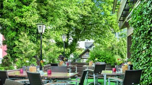 An outdoor seating area with tables and chairs, surrounded by lush greenery and a building with a sign reading "Hacker Partner". - Hotel Thüringenschanze (Oberhof)