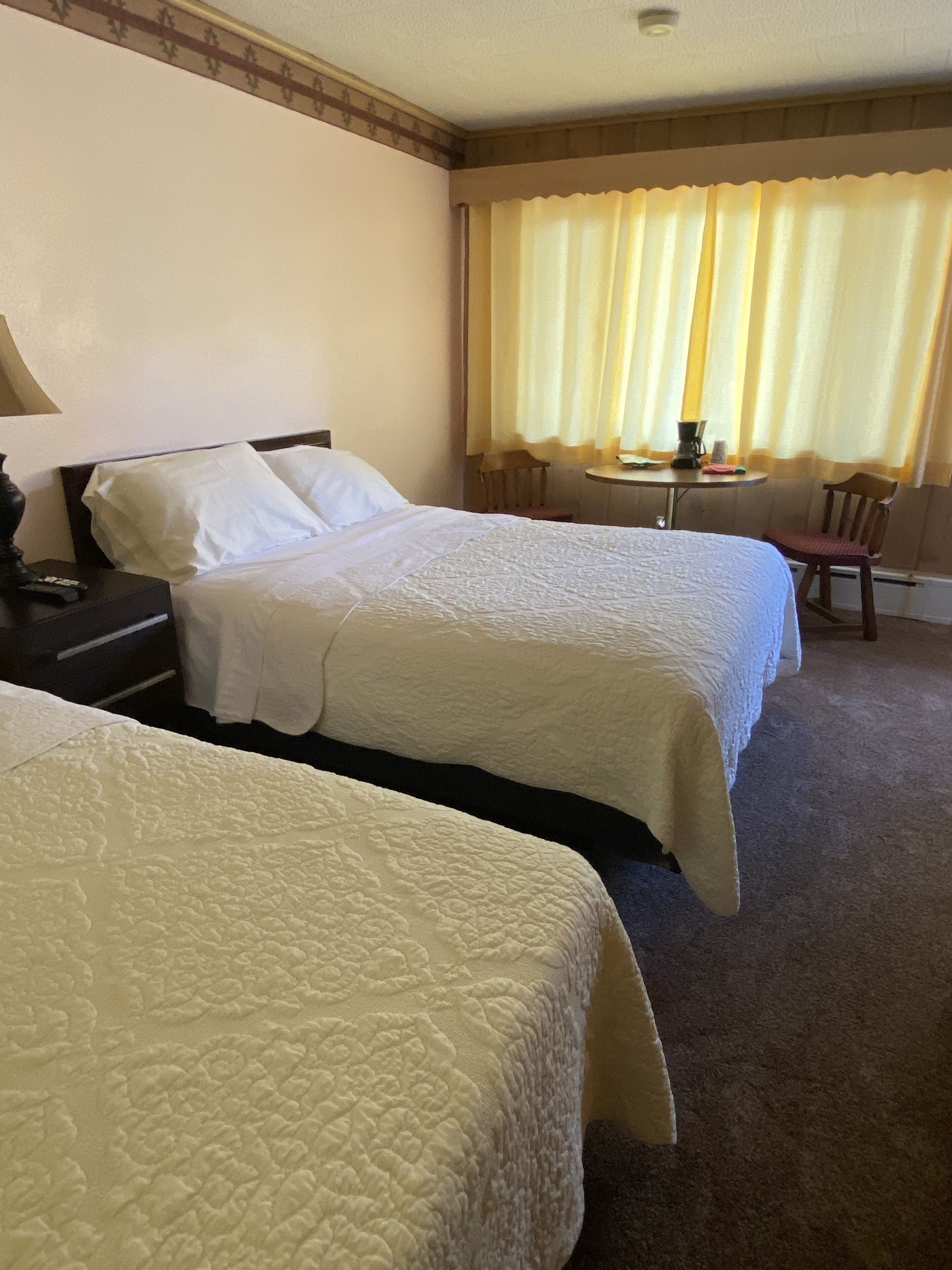 A hotel room with two beds, a small table with a lamp, and a window with curtains.