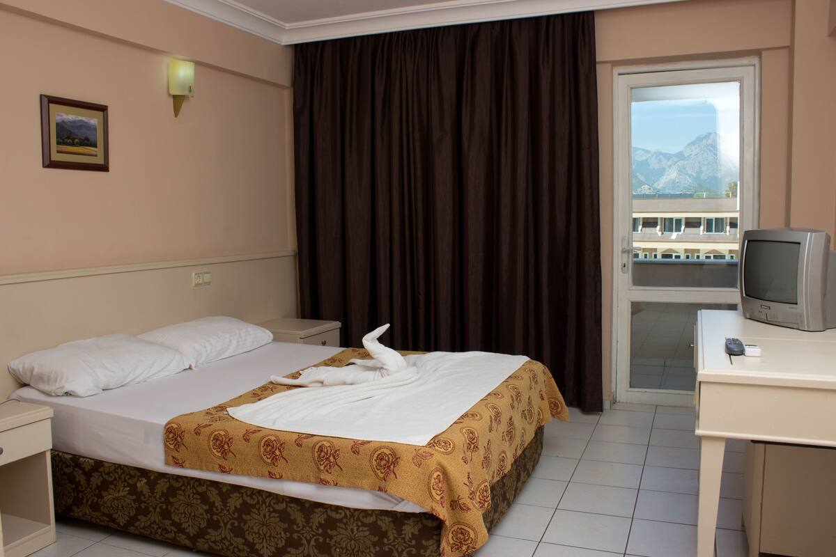 Quarto de hotel com cama, escrivaninha com televisão, janela com vista e porta que dá para uma varanda.
