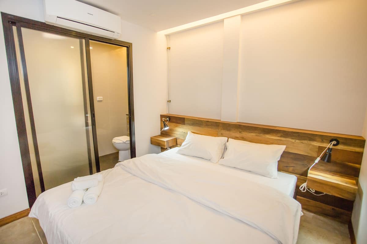 A hotel room with a bed, a wooden headboard, a nightstand, a bathroom with a toilet, and a sliding door.