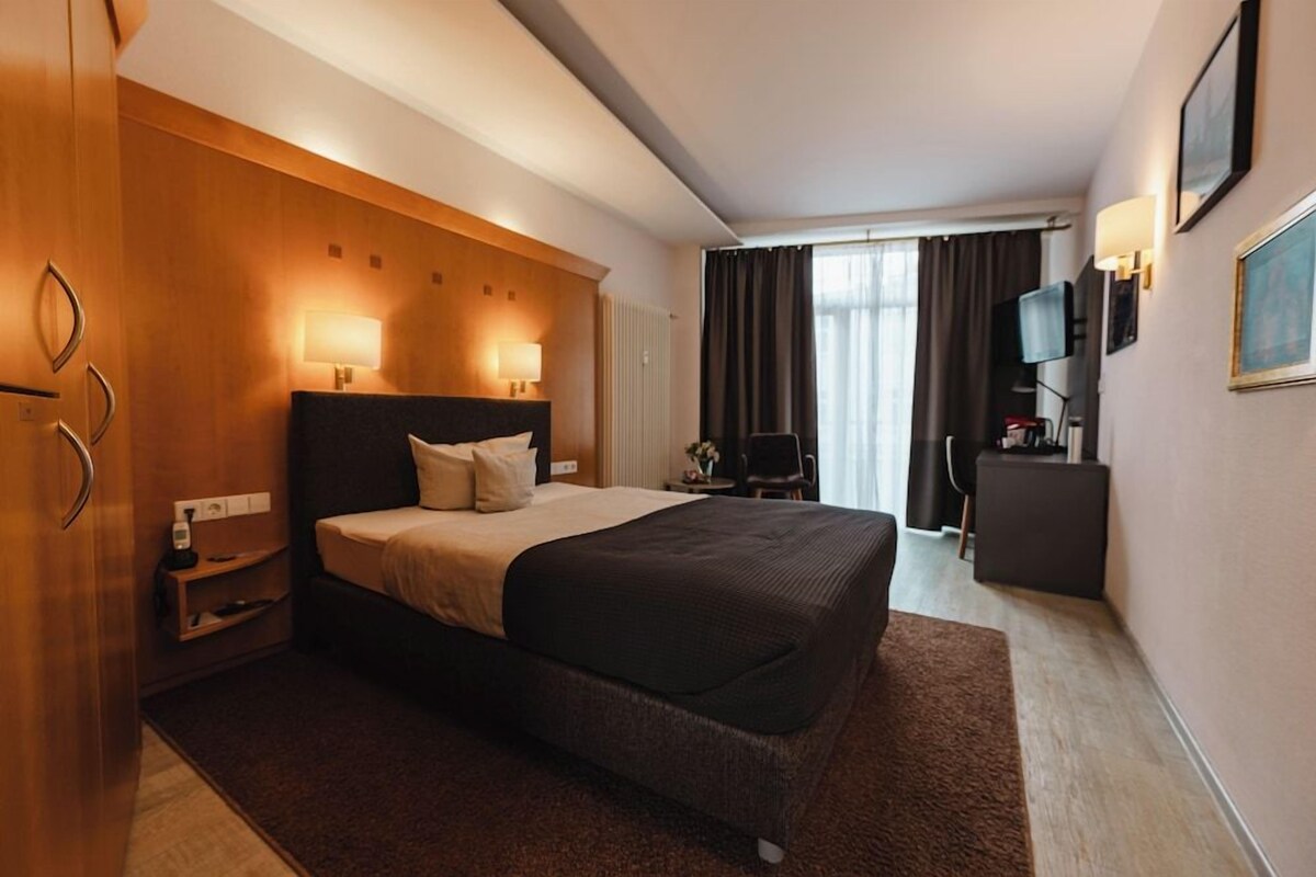 A hotel room with a large bed, bedside lamps, a desk with a chair, a television, and a window with curtains.