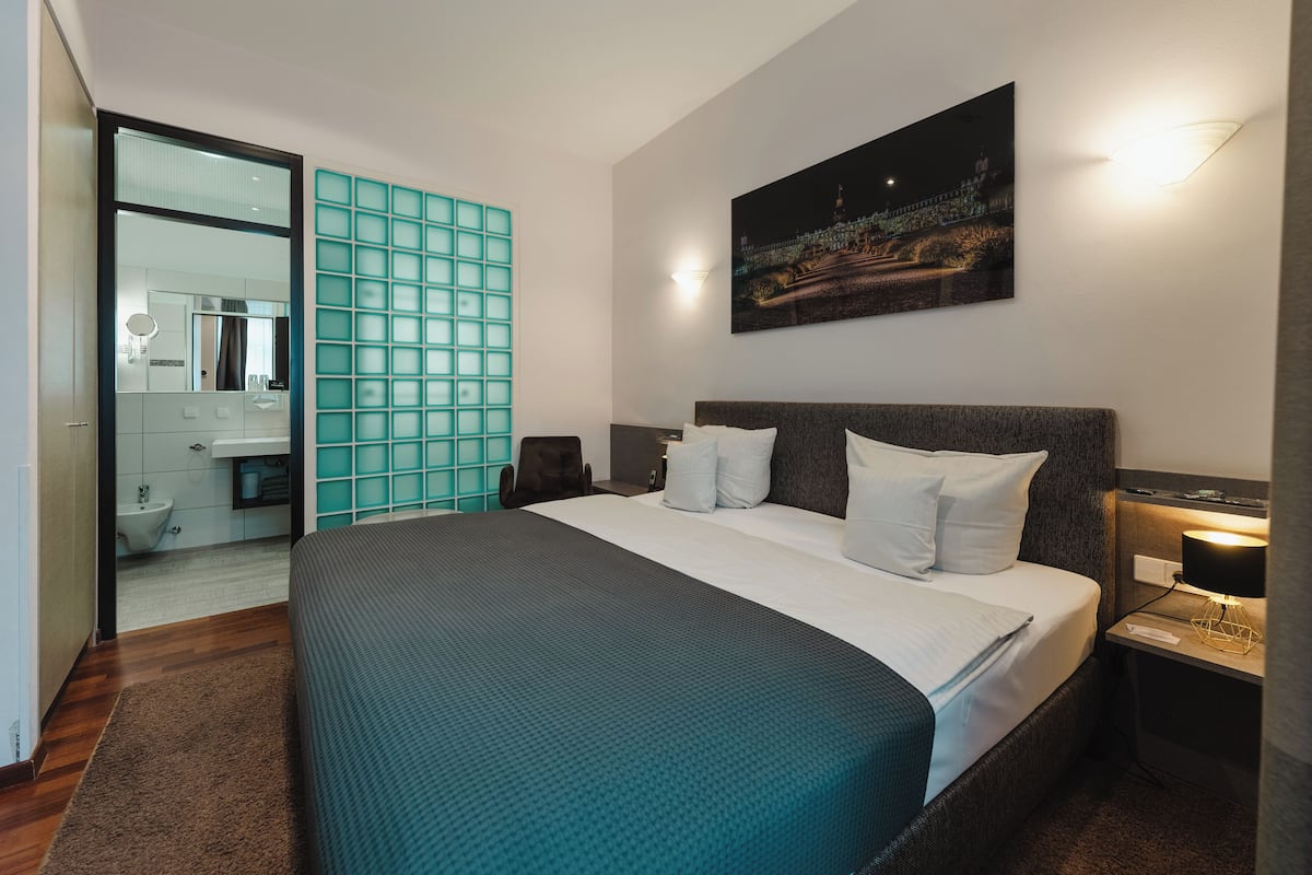 A modern hotel room with a large bed, a nightstand with a lamp, and a wall-mounted painting.