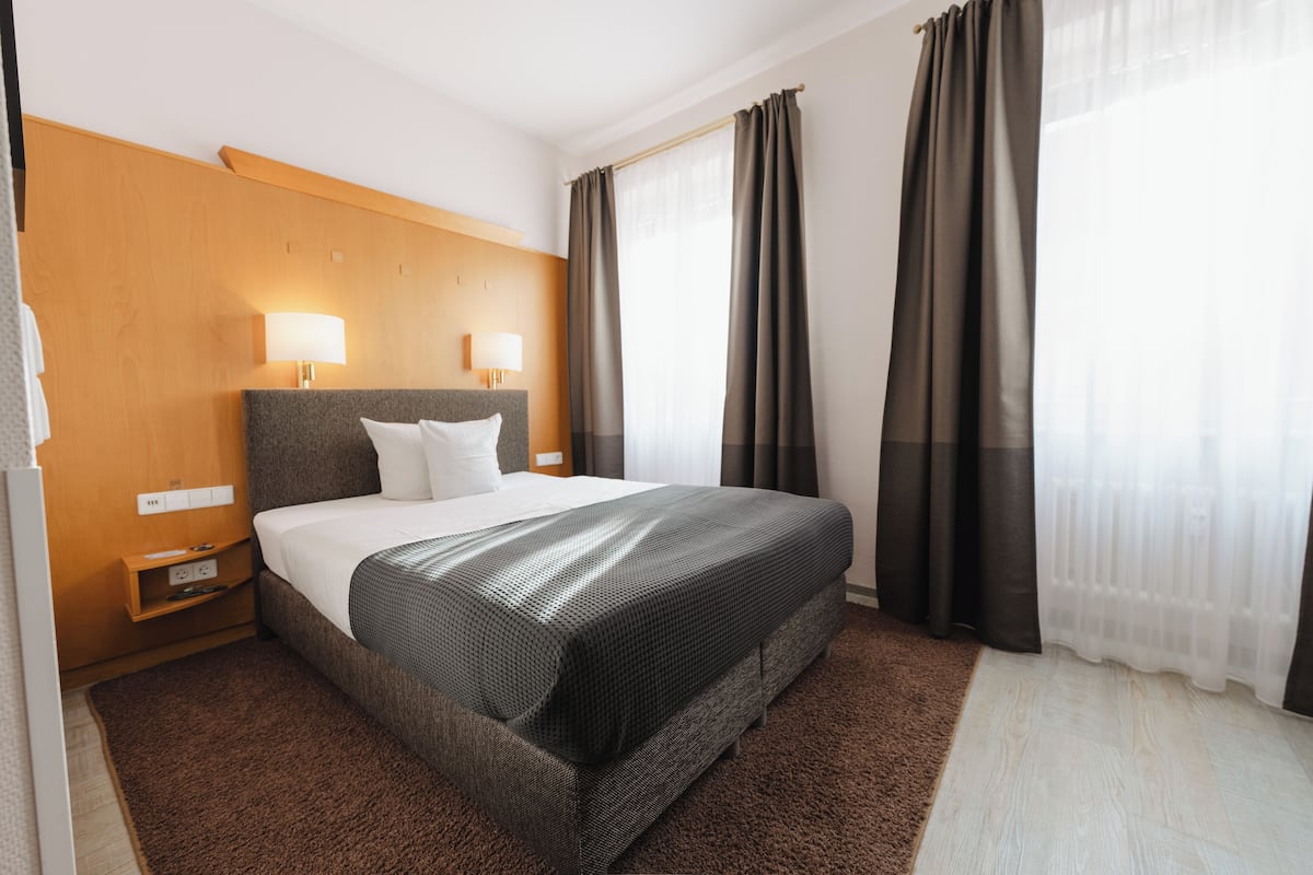 A hotel room with a large bed, two bedside lamps, a wooden headboard, and a view of the outdoors through sheer curtains.