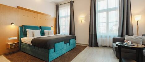A modern hotel room with a large bed, a sofa, a small table, and a window with curtains.