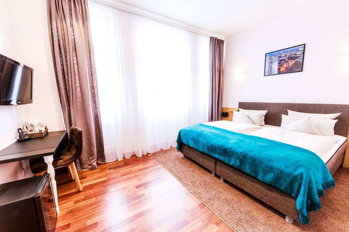 A hotel room with a bed, a desk, a chair, a TV, and a window with curtains.
