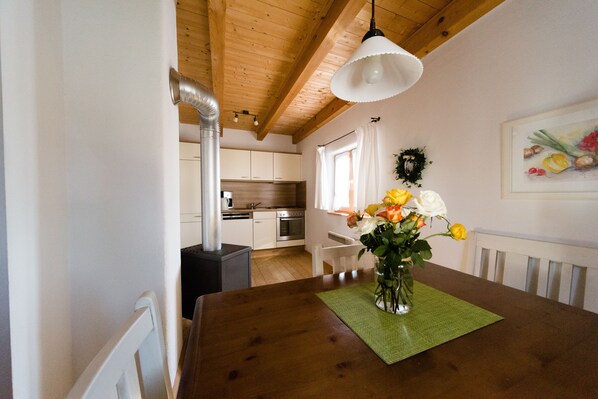 Un comedor con una mesa de madera, un florero y vista a una cocina con gabinetes blancos y techo de madera.