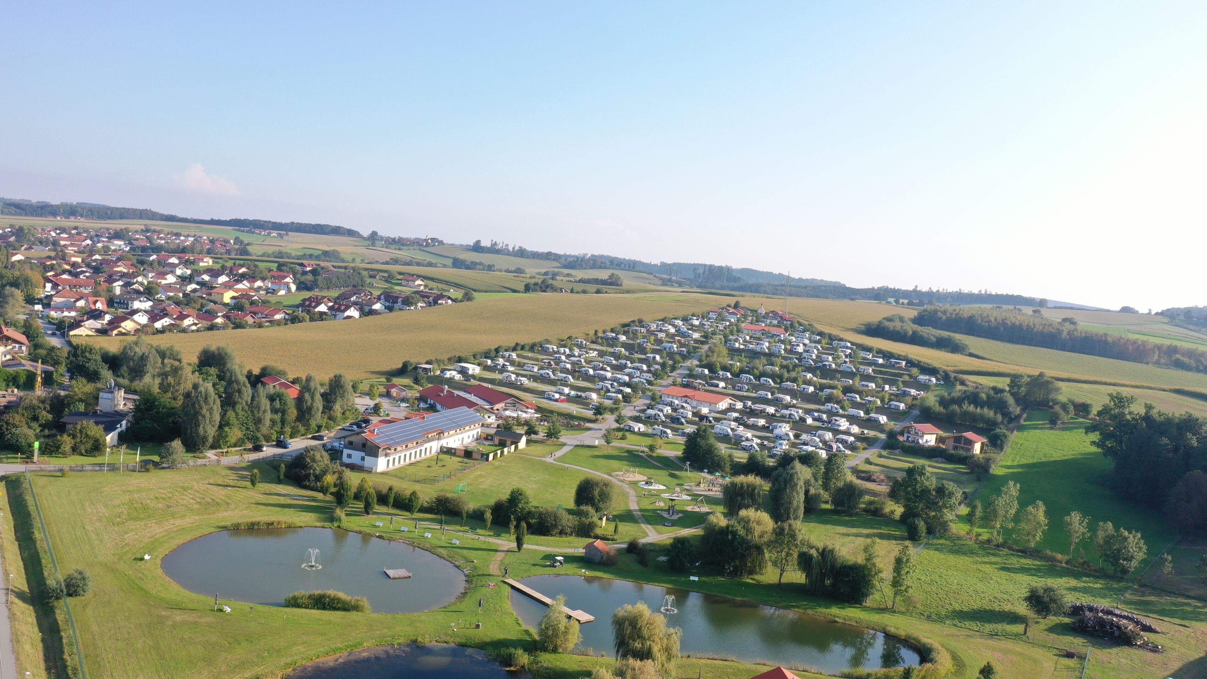 A large campground with numerous RVs and tents, surrounded by green fields and a few buildings.