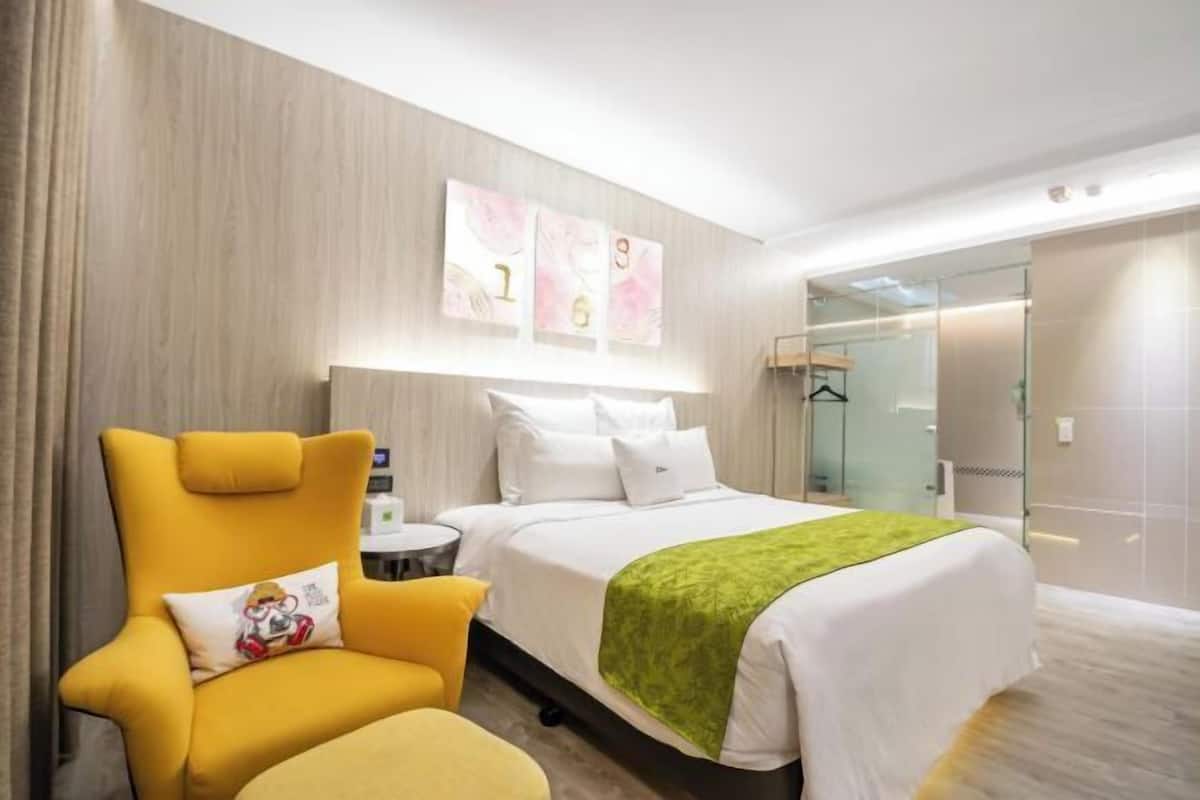A hotel room with a bed, a chair, a bedside table, and a glass shower area.