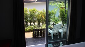 Un balcon avec des chaises en plastique blanches, une porte vitrée et une vue sur un jardin où poussent différentes plantes, ainsi qu’un sol carrelé.