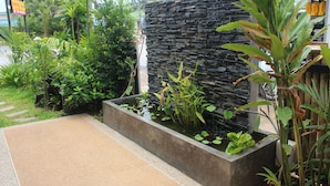 Un espace paysager aménagé en jardin, avec un mur en pierre, diverses plantes et un bac en béton contenant de l’eau et des nénuphars.