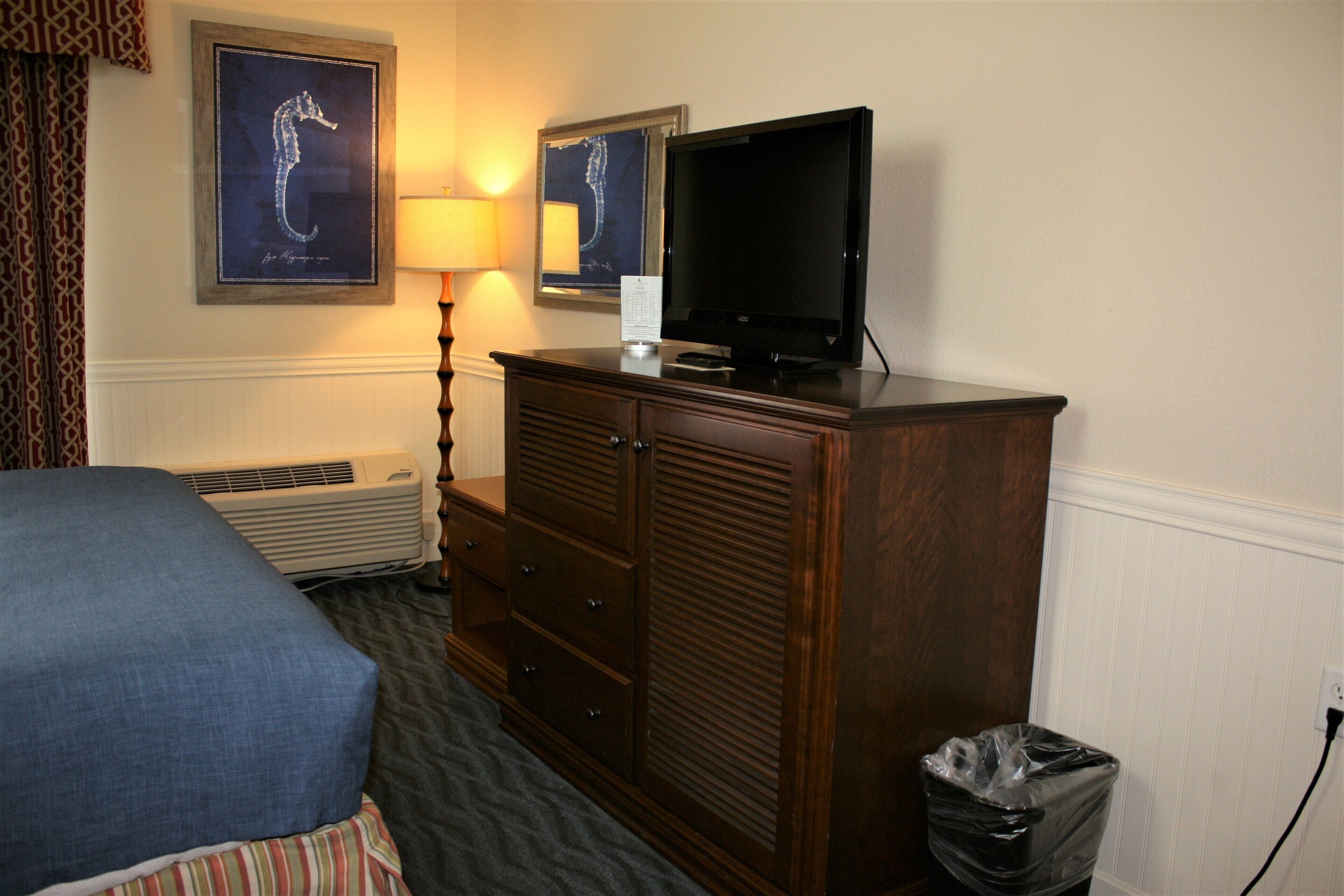 A hotel room with a bed, a wooden dresser, a television, and a wall-mounted air conditioner.
