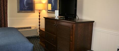 A hotel room with a bed, a wooden dresser, a television, and a wall-mounted air conditioner.