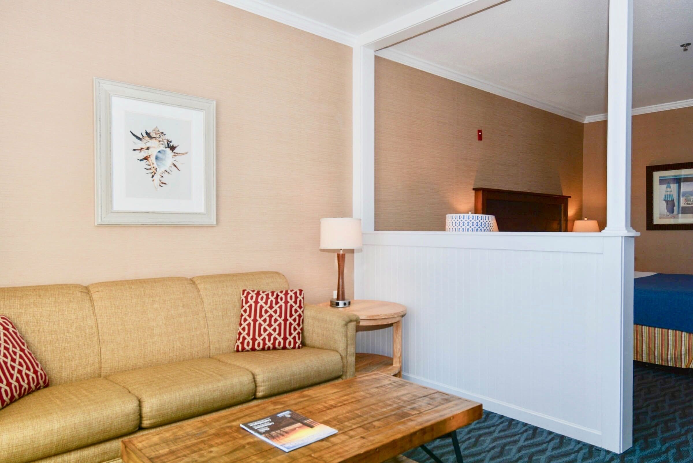 A hotel room with a beige sofa, a wooden coffee table, a small side table with a lamp, and a framed picture on the wall.