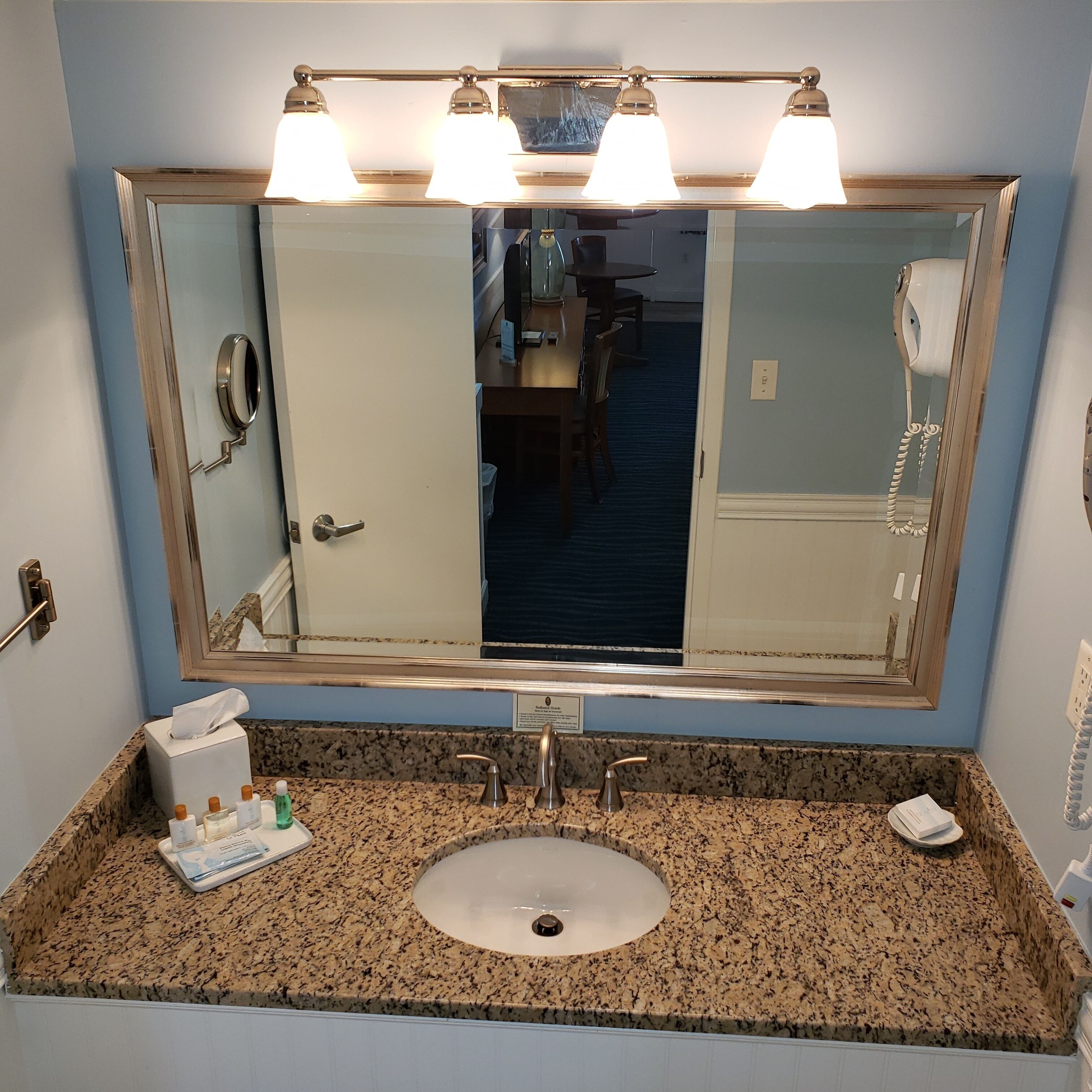 A bathroom with a granite countertop, a sink, a mirror with three lights, and a hairdryer.