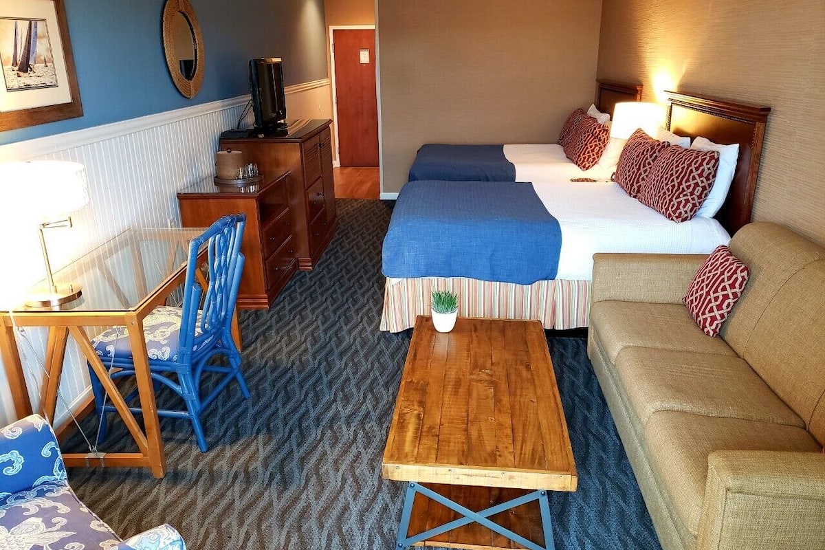 A hotel room with a bed, a sofa, a wooden table, a chair, and a small table with a plant.