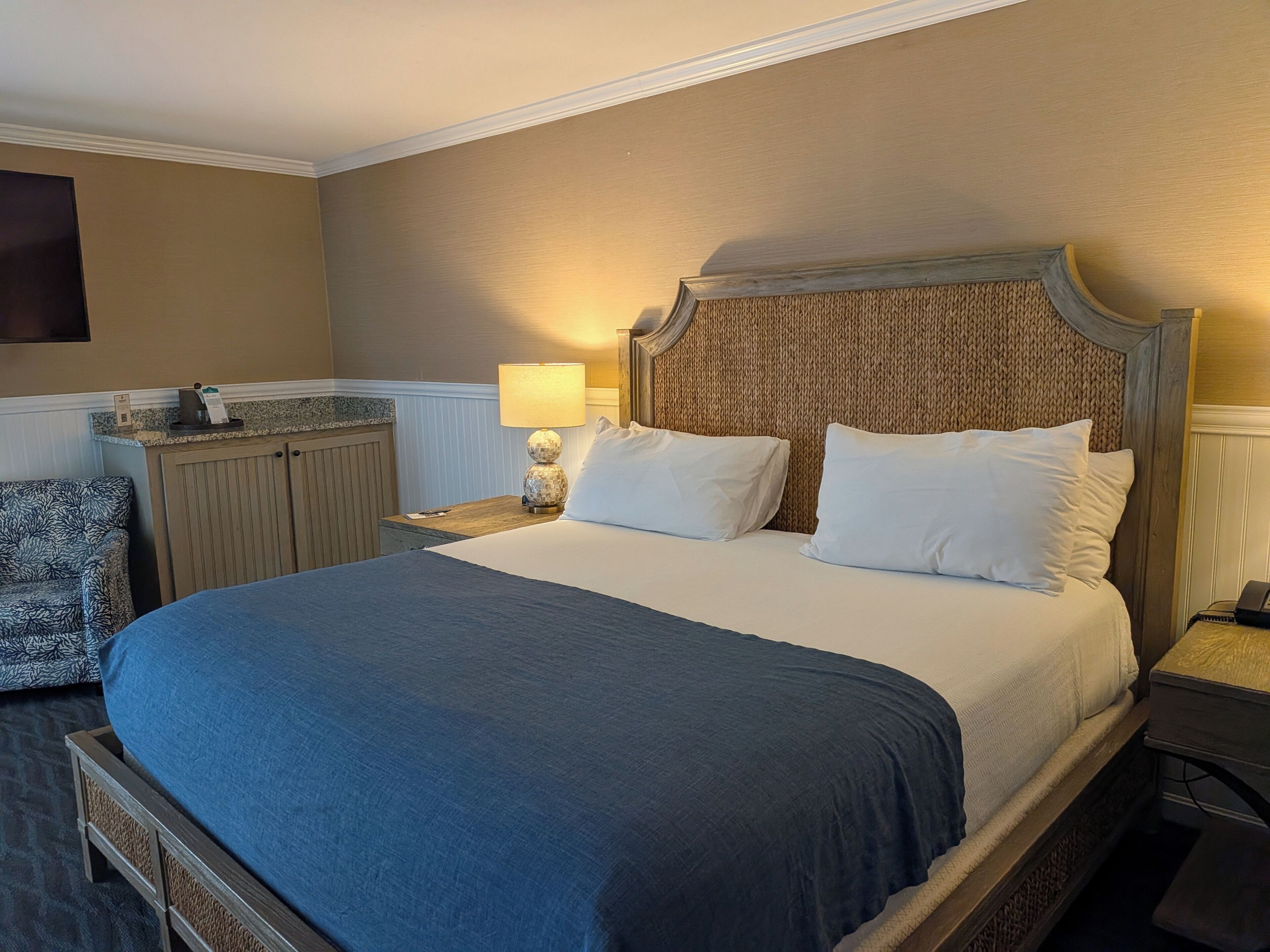 Ein Hotelzimmer mit einem großen Bett, einem Nachttisch mit Lampe, einem Holzkopfende und einem gemusterten Sessel.