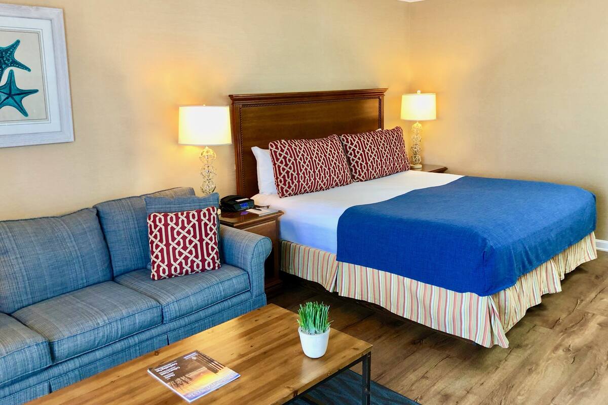 A hotel room with a bed, a blue sofa, a wooden coffee table, and a framed picture on the wall.