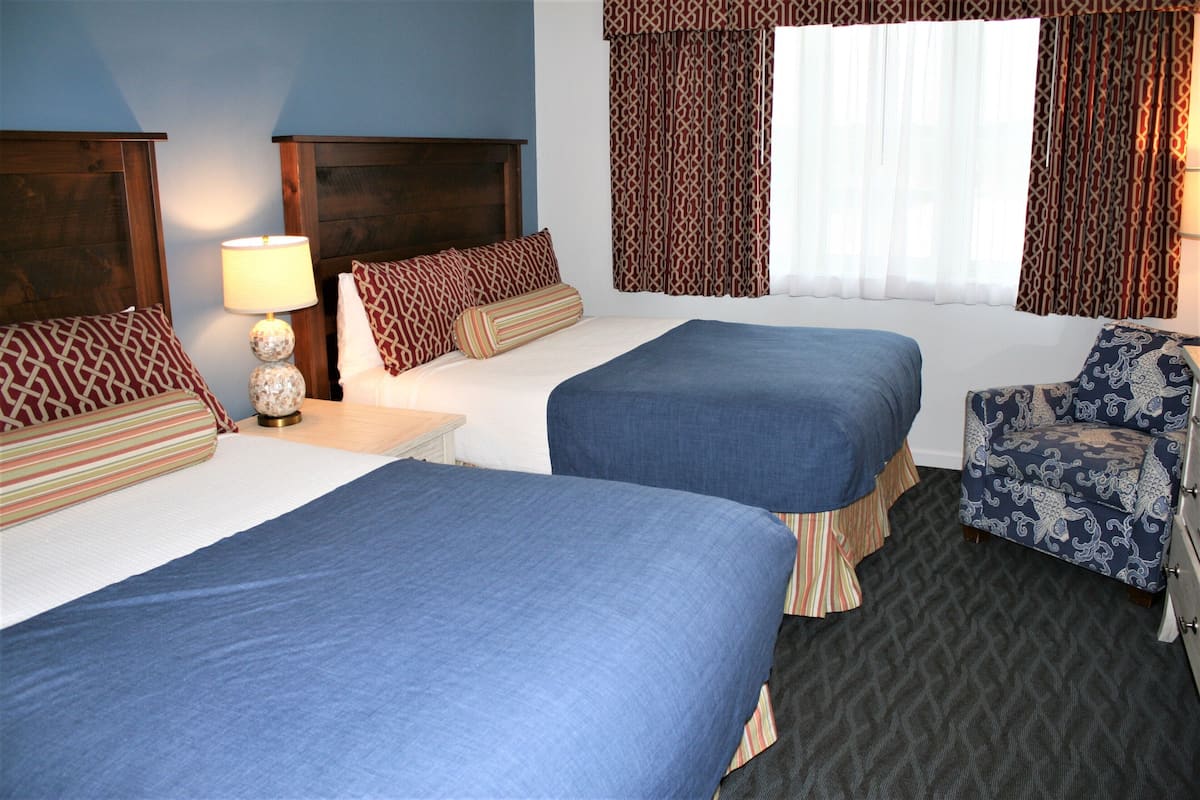 A hotel room with two beds, a nightstand with a lamp, a chair, and patterned curtains.