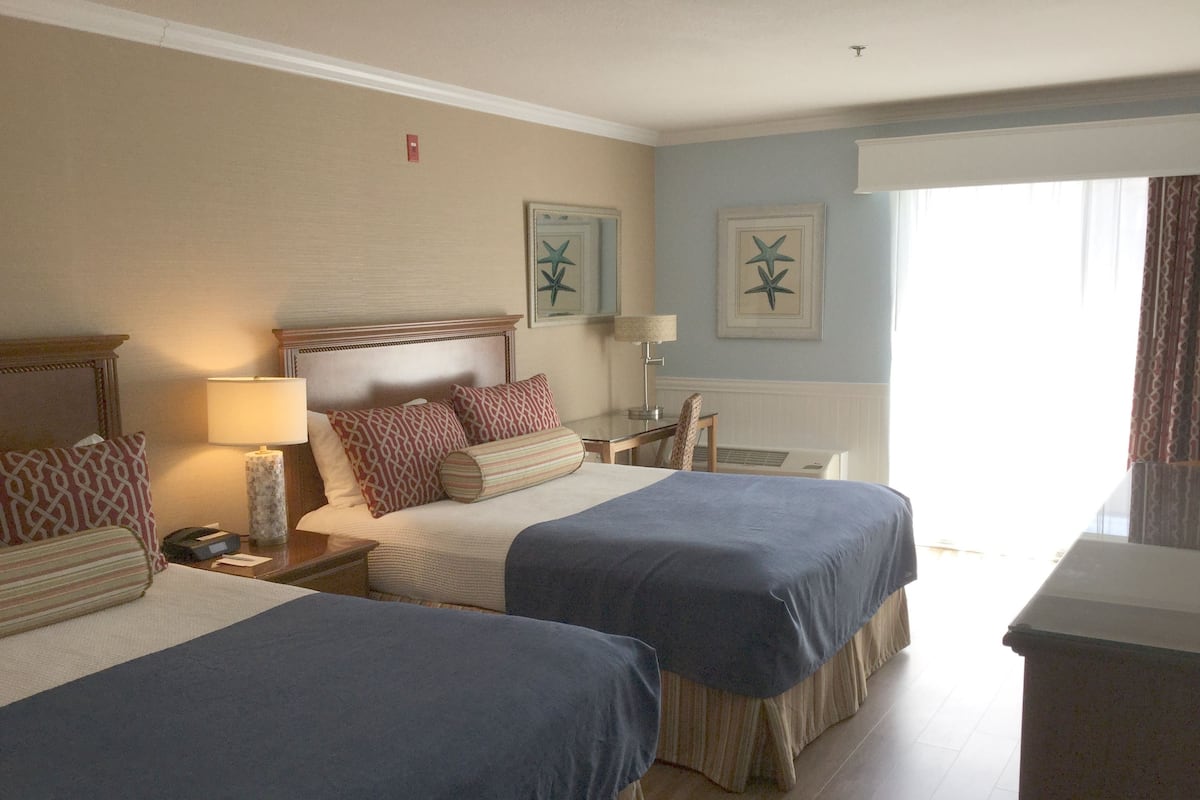 A hotel room with two beds, a nightstand with a lamp, a desk, and a window with curtains.