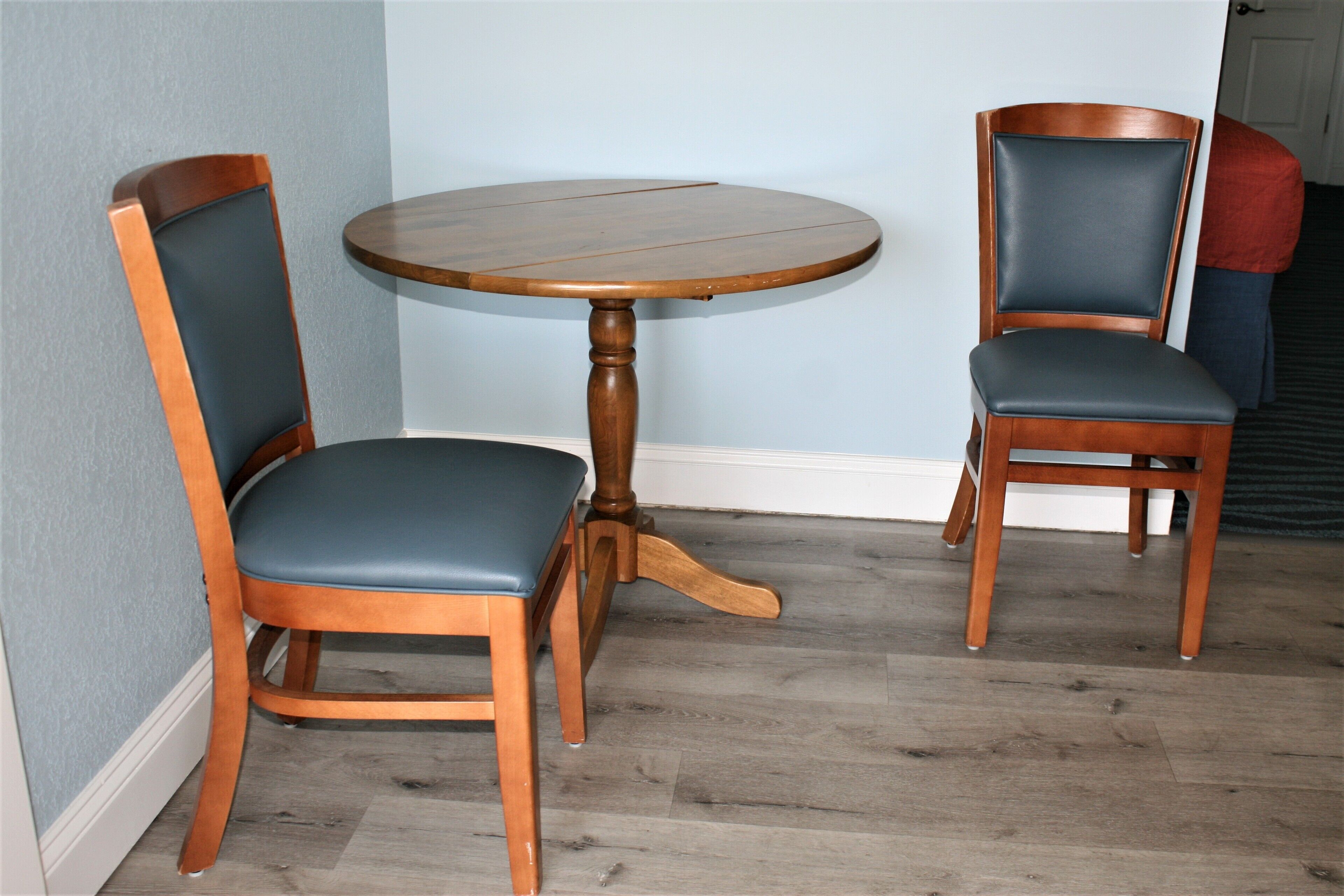 A round wooden table with two chairs, each with a cushioned seat and backrest, positioned in a corner of a room with wooden flooring and light blue walls.