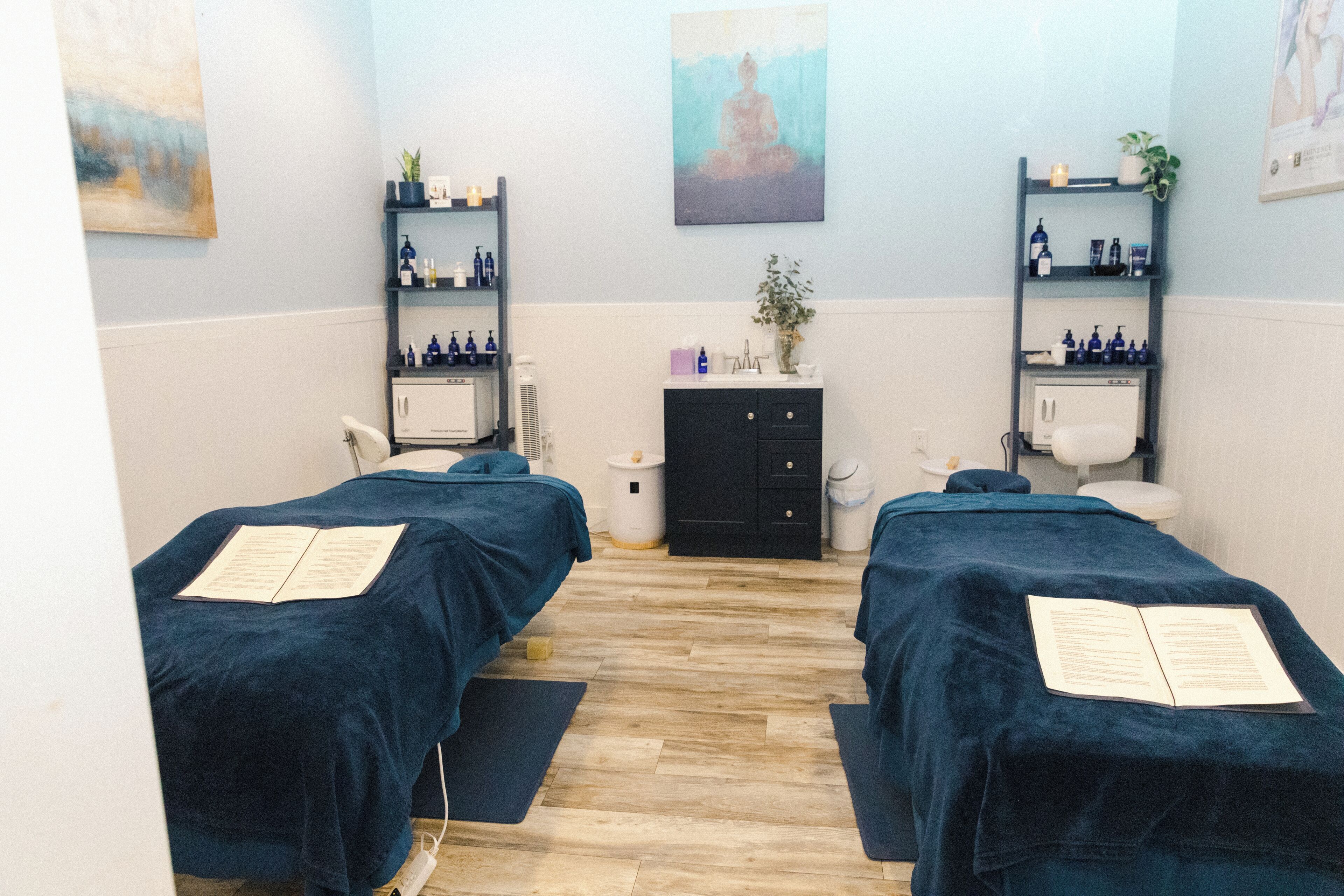 Un cuarto de spa con dos camillas de masaje, un mueble bajo, repisas con botellas y un cuadro en la pared.