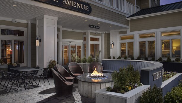 Exterior de un hotel con un letrero que dice "THE AVENUE", mobiliario exterior con sillas y un brasero, y un jardín ajardinado.