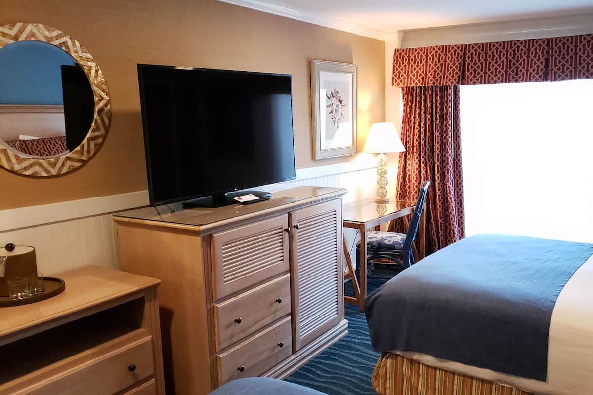 A hotel room with a flat-screen TV, a desk with a lamp, a chair, and a bed.