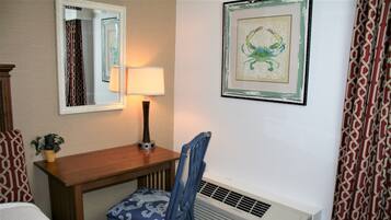 A hotel room with a wooden desk, a chair with a patterned cushion, a wall-mounted lamp, a framed artwork, and a radiator.