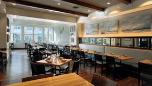 Un restaurante espacioso con techos altos, vigas de madera y ventanas amplias que dejan entrar luz natural. Las mesas están ordenadas con sillas, y se exhiben obras de arte en las paredes.