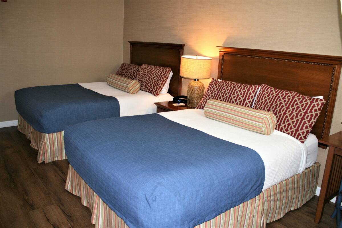 A hotel room with two beds, each with a blue bedspread and striped skirted beddings. There is a wooden headboard, a nightstand with a lamp, and a wooden nightstand with a phone.
