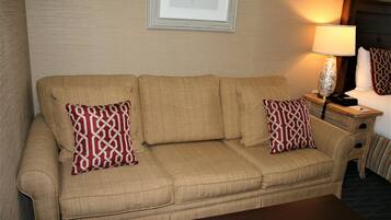 A beige sofa with patterned pillows, a wooden nightstand with a lamp, and a framed picture on the wall.