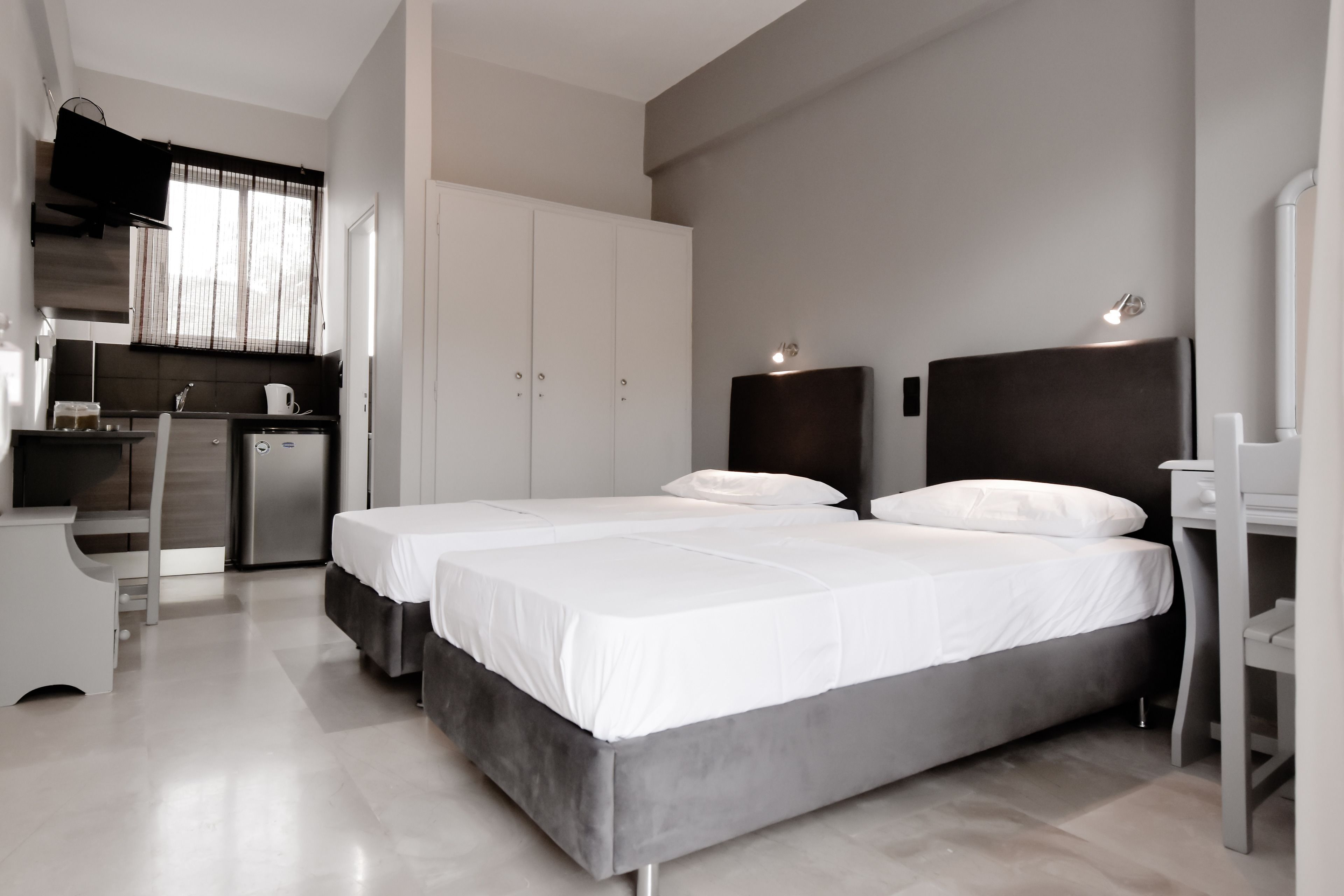 A modern hotel room with two beds, a desk, a kitchenette, and a large wardrobe.