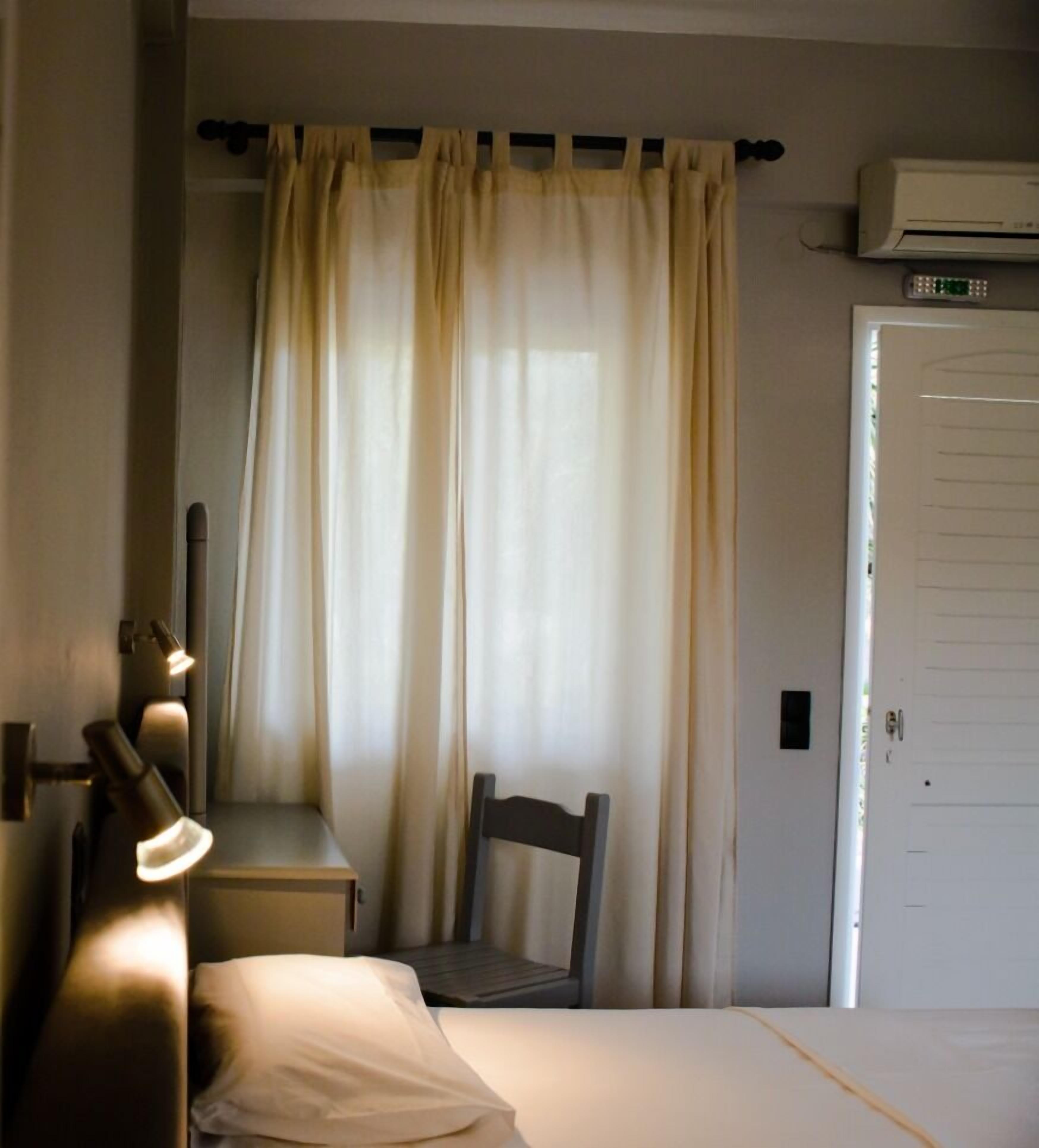 Un dormitorio con cama, una silla, escritorio con lámpara, ventana con cortinas y aire acondicionado.