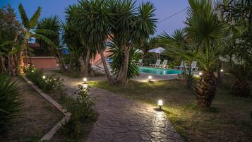 Un espacio exterior nocturno con piscina, un sendero de piedra y palmeras.