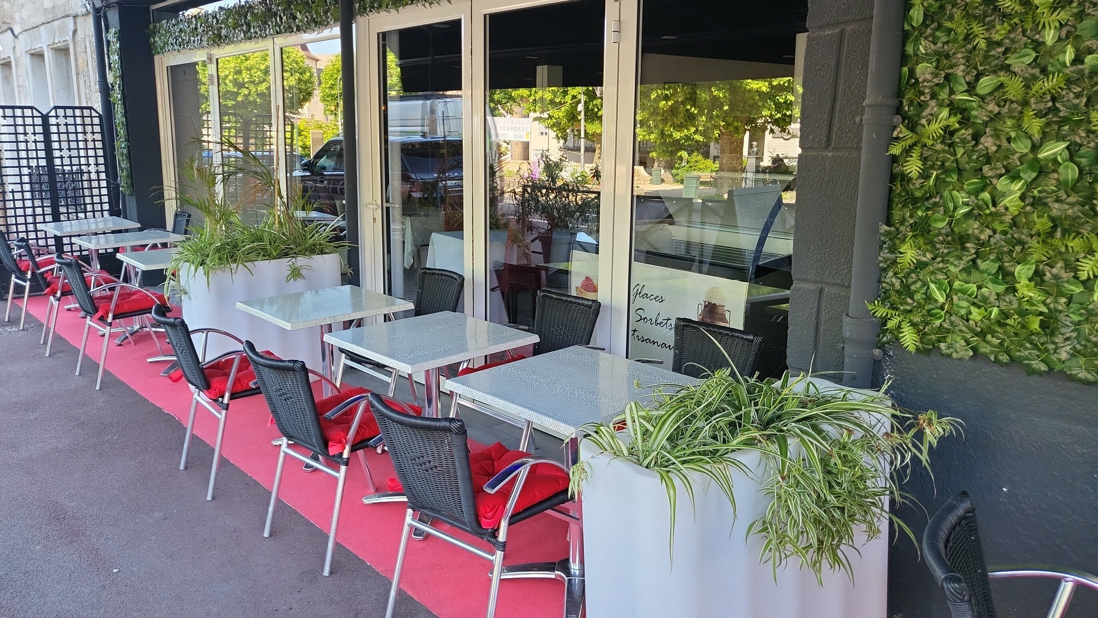 Un café al aire libre con mesas y sillas de vidrio, plantas en macetas y vista a la calle.