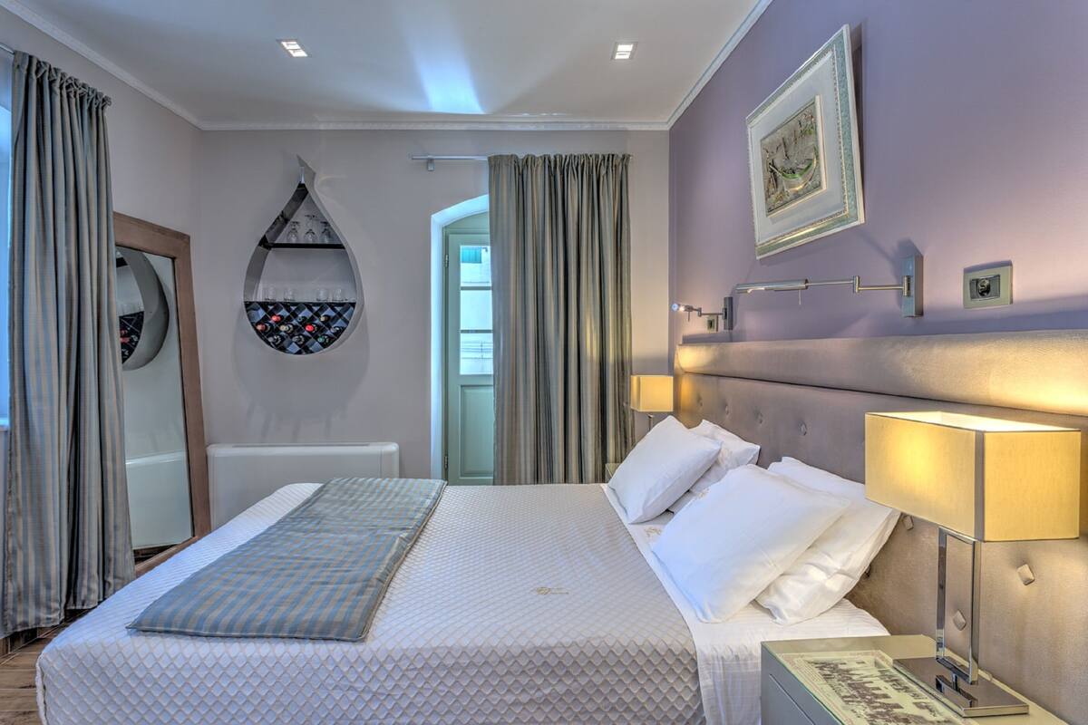 Quarto de hotel com cama grande, luminárias laterais, espelho e vista para o exterior.