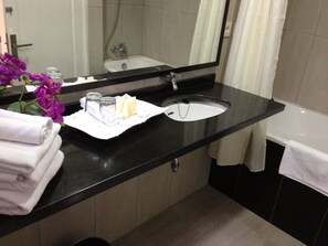 A modern bathroom with a black countertop, white sink, and a large mirror. There are towels stacked on the counter and a small plant with purple flowers.