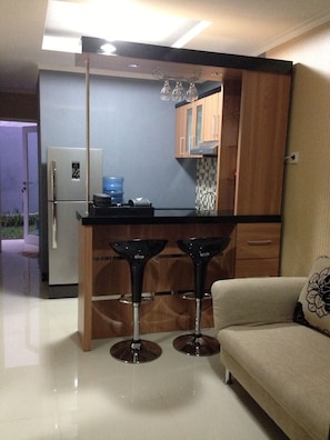 A compact kitchen with a refrigerator, a water dispenser, and a coffee machine. There are two black bar stools and a beige sofa with a floral pillow.