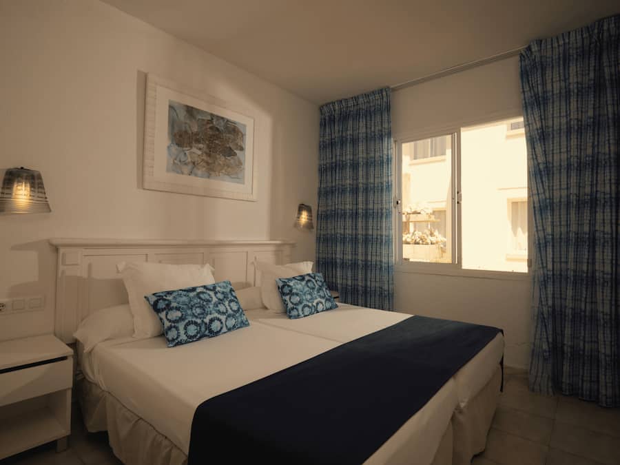 Un dormitorio con cama, mesitas de noche, lámpara de pared, ventana con cortinas y un cuadro enmarcado.