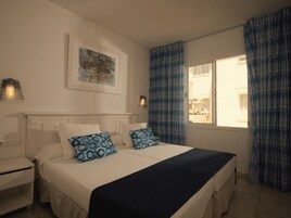 Un dormitorio con cama, mesitas de noche, lámpara de pared, ventana con cortinas y un cuadro enmarcado.