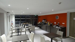 Un comedor moderno con sillas y mesas blancas, una barra de cocina con electrodomésticos y un ventanal con vista a coches estacionados.
