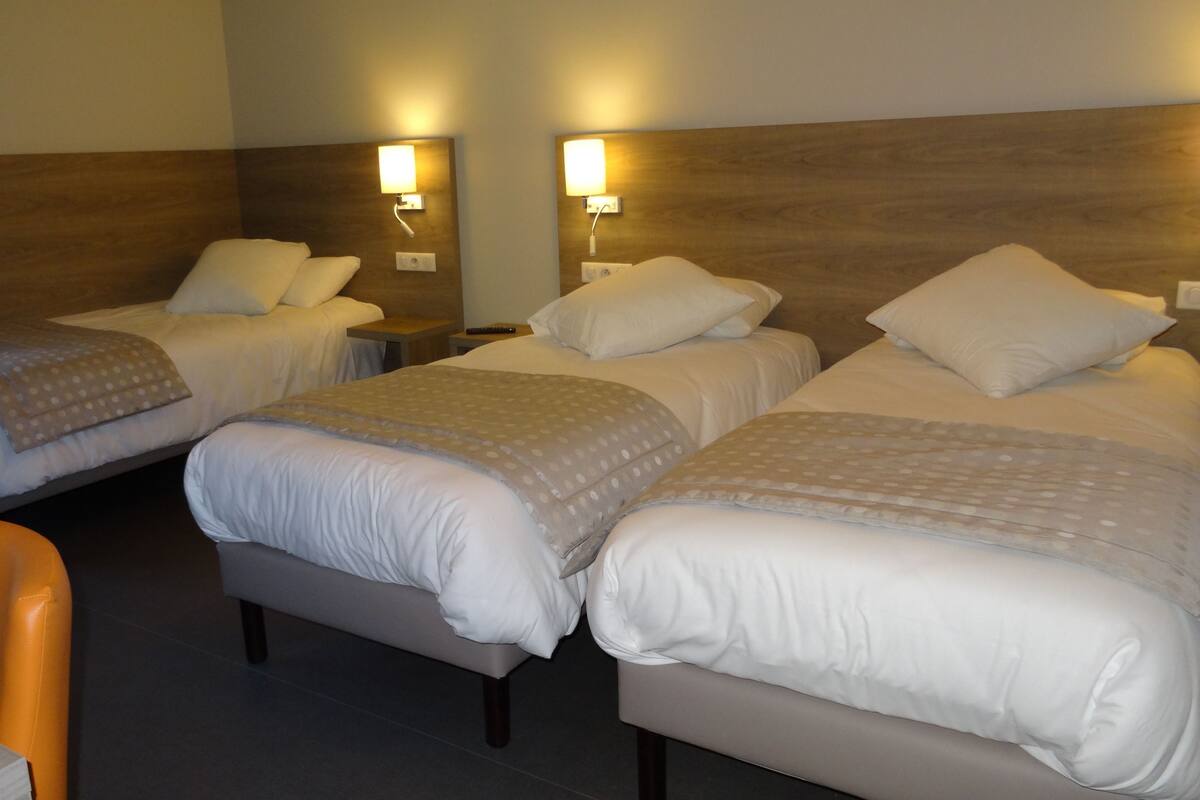 A hotel room with two single beds and one double bed, each with white bedding and a patterned throw. There are bedside lamps and a small table with a chair.