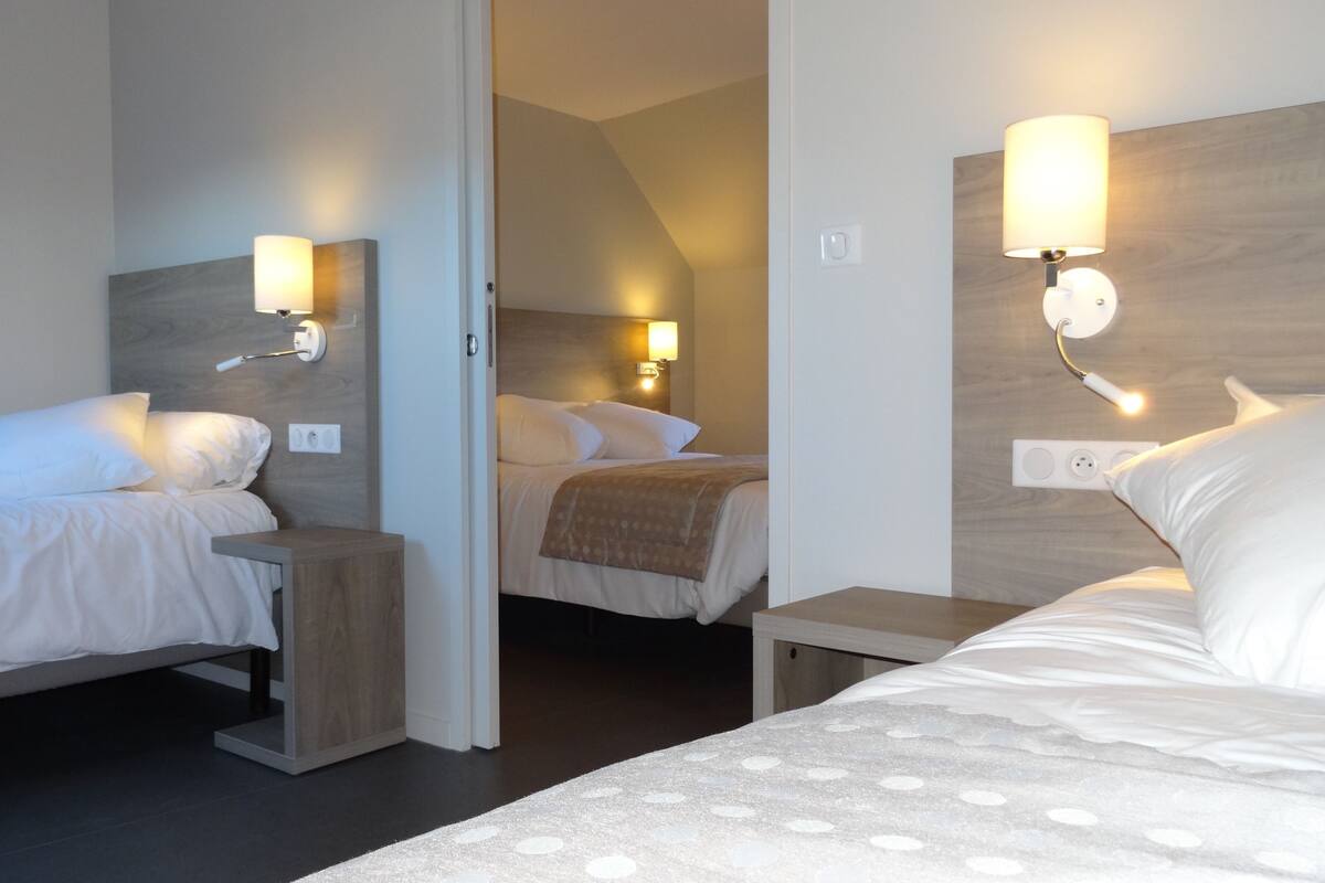 A hotel room with three beds, each with a nightstand and wall-mounted reading lamp.